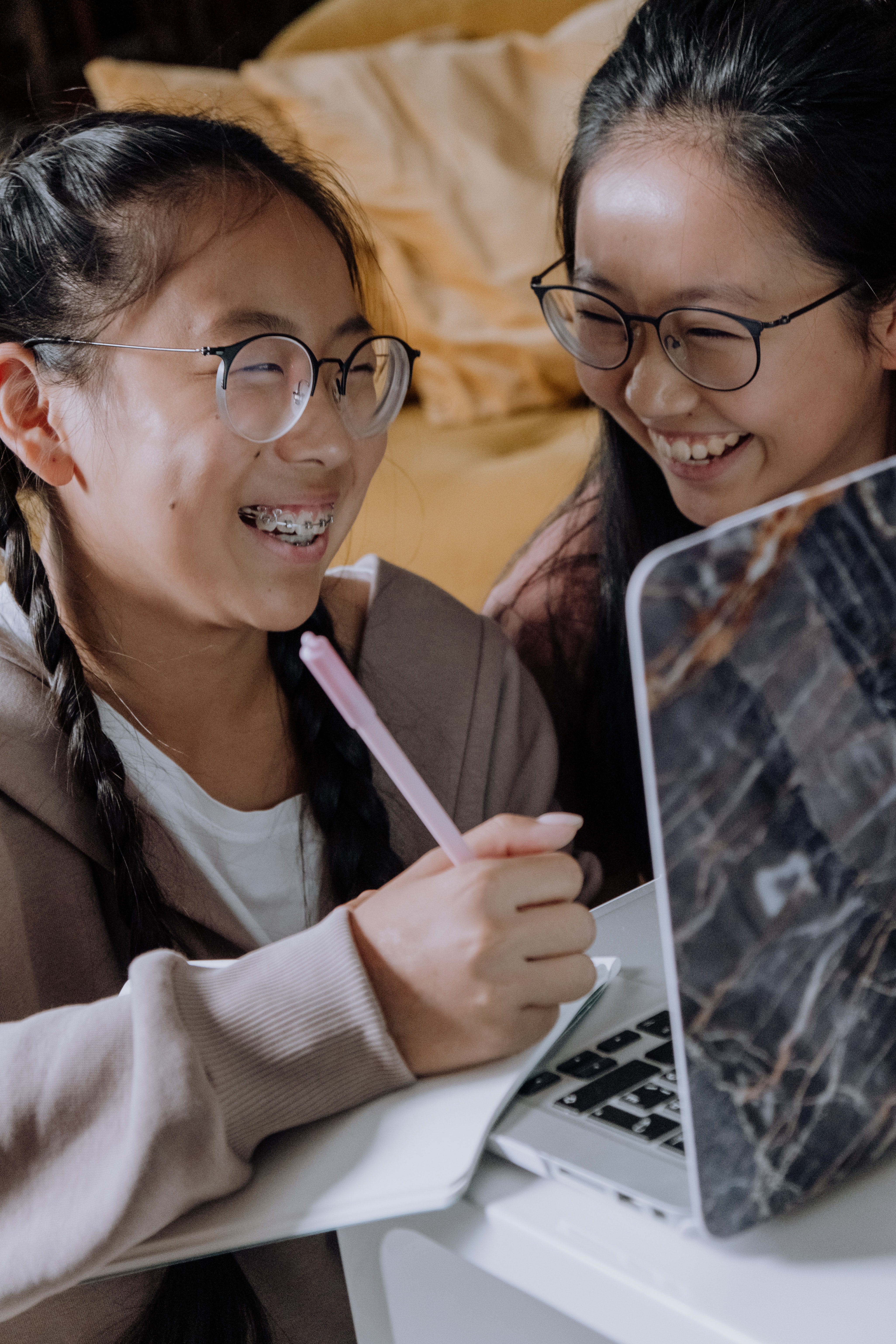 Dos jóvenes asiáticas con gafas riendo mientras hacen deberes en una libreta y un ordenador