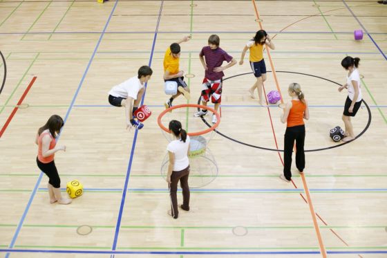 Se visualiza un gimnasio escolar con alumnos jugando con unos dados grandes en la pista de baloncesto y la profesora de espaldas.