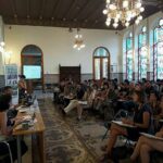 se puede apreciar a un gran número de personas sentados en una sala. hay tres personas en una gran mesa, hablando o exponiendo cosas al resto, que son el público
