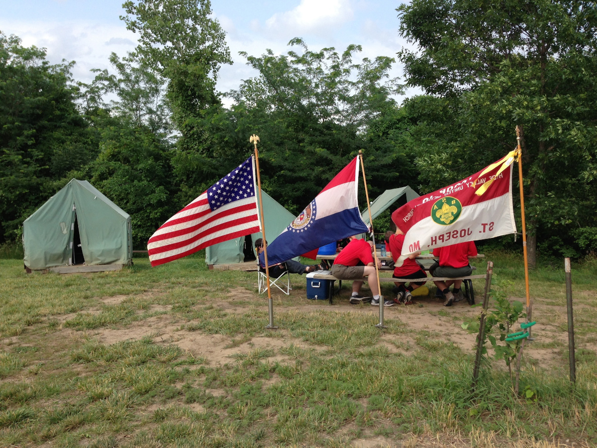 fotografía de un campamento con tres tiendas de campaña y cerca de ellas niños y niñas sentados en mesas de camping y también banderas de distintos países
