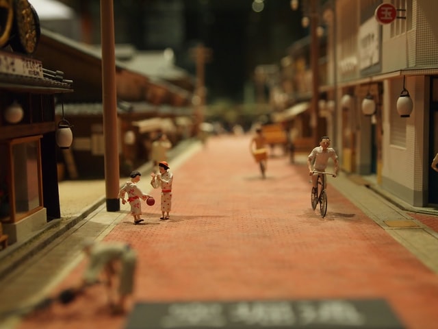 La imagen muestra un diorama de una calle de una población asiática. Una maqueta de una calle central con casas a ambos lados. En el centro se ve un hombre en bicicleta y dos niños jugando.