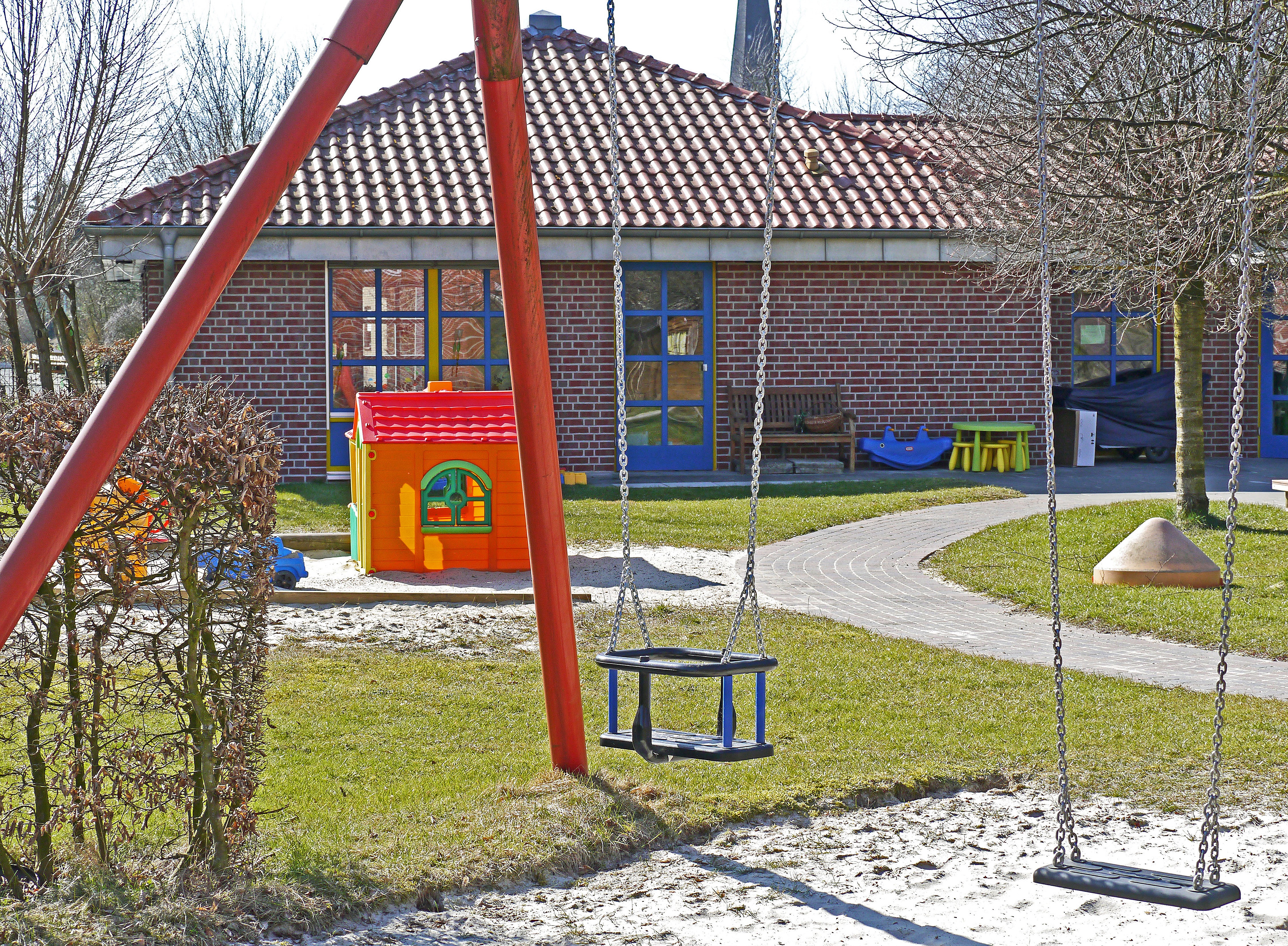 La imagen muestra una zona de juegos con césped, un columpio y una caseta de juguete.