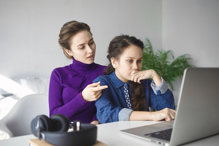 Chica mirando atentamente la pantalla de un ordenador portátil, mientras otra mujer, detrás de ella, parece darle instrucciones