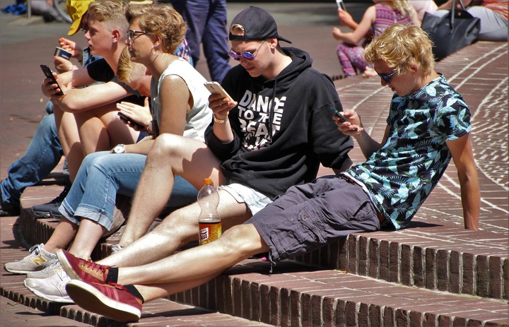 Grupo de varias personas, sentadas al aire libre, mirando sus dispositivos móviles.