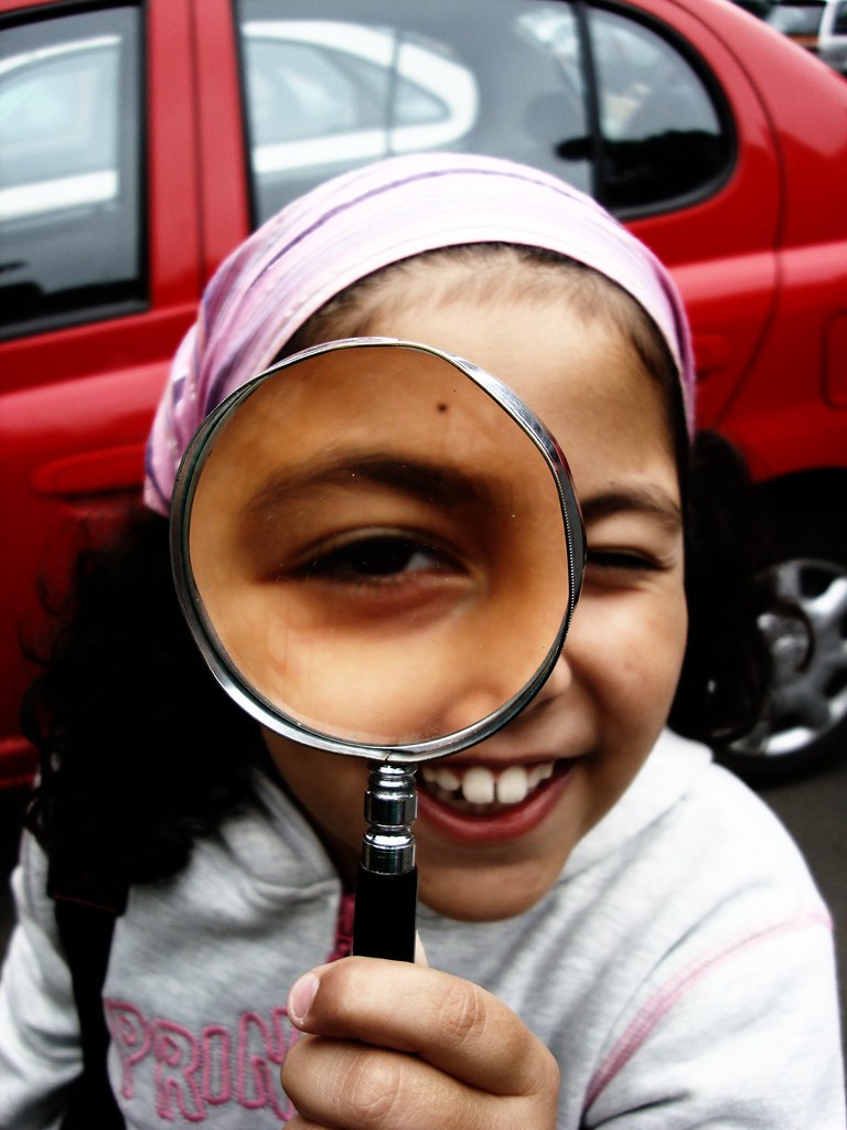 La imagen muestra una niña con una lupa pegada a su ojo derecho.