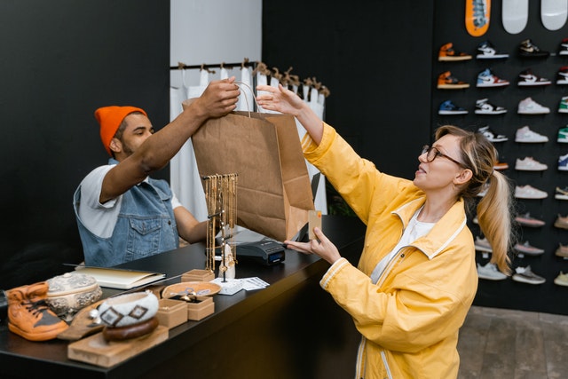 La imagen muestra a un dependiente de ventas entregando una bolsa a un cliente y en el fondo de la imagen se aprecian estanterías de zapatillas deportivas. 