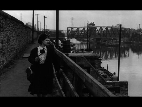 La imagen en blanco y negro muestra a una mujer cruzando un puente en una ciudad.