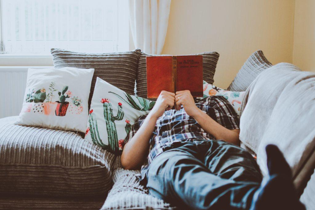 La imagen muestra una persona tumbada en un sofá y leyendo un libro.