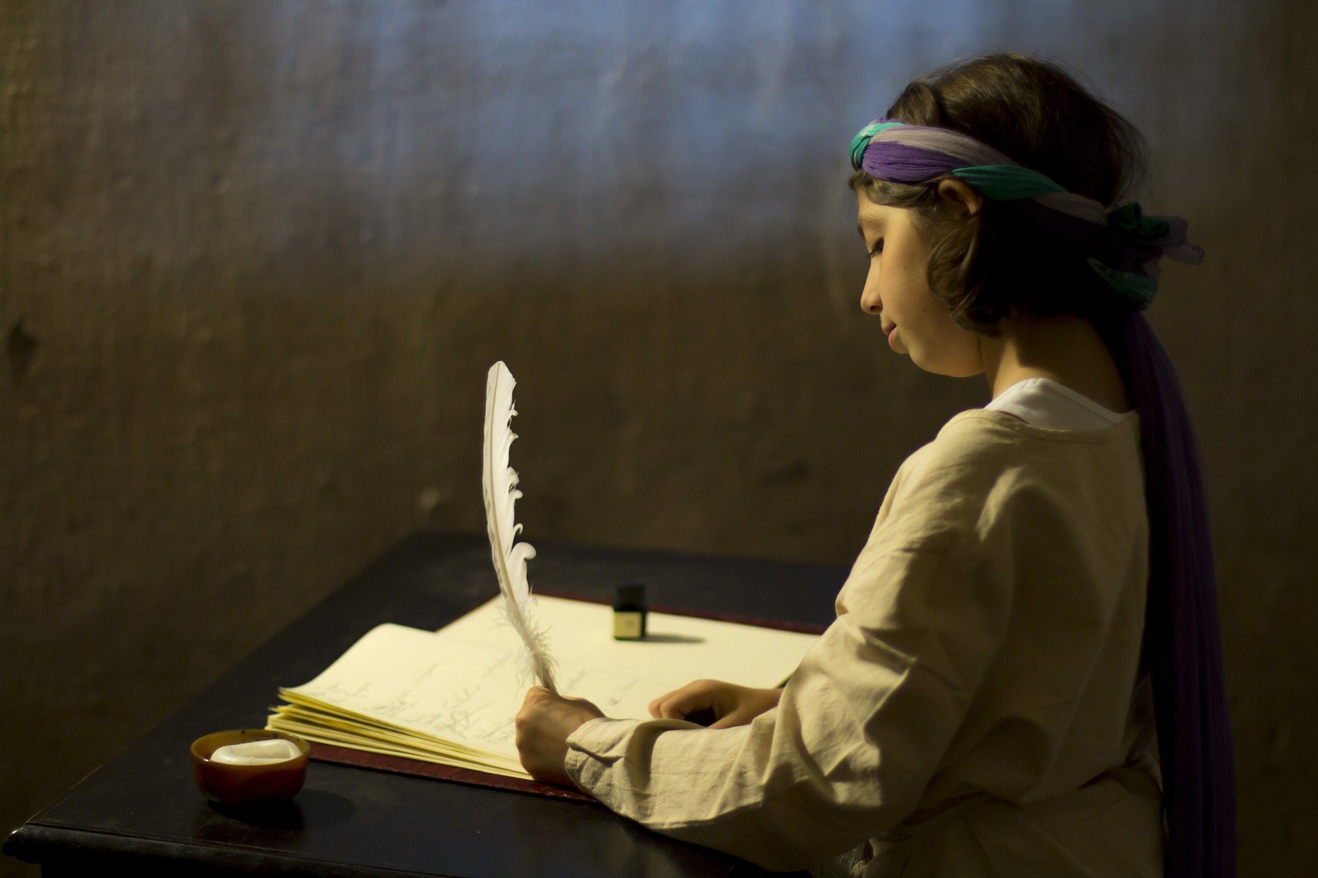 En la penumbra, un niño o una niña escribe con pluma en un cuaderno en blanco.