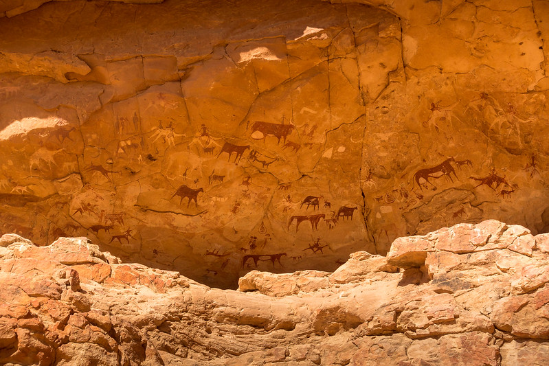 La imagen muestra pinturas rupestres de color rojo y blanco sobre un muro de piedra en las que aparecen animales similares a caballos y personas montando a caballo