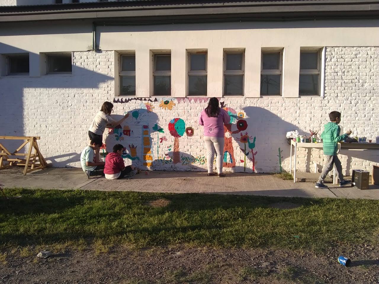 La imagen muestra unos niños y niñas pintando murales sobre una pared blanca