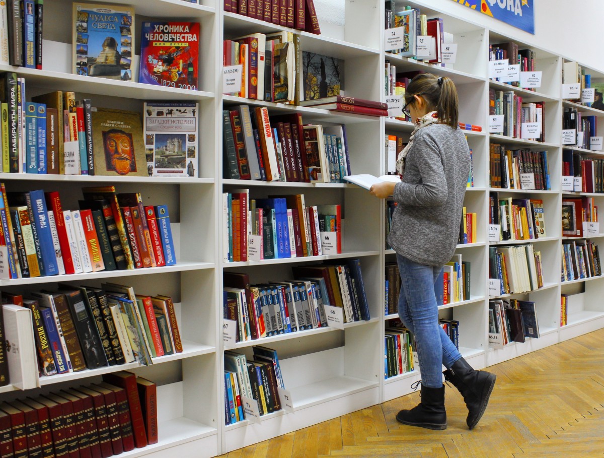Imagen de una bilbioteca, con estanterías blancas llenas de libros y una mujer de espalda ojeando un libro abierto en sus manos.