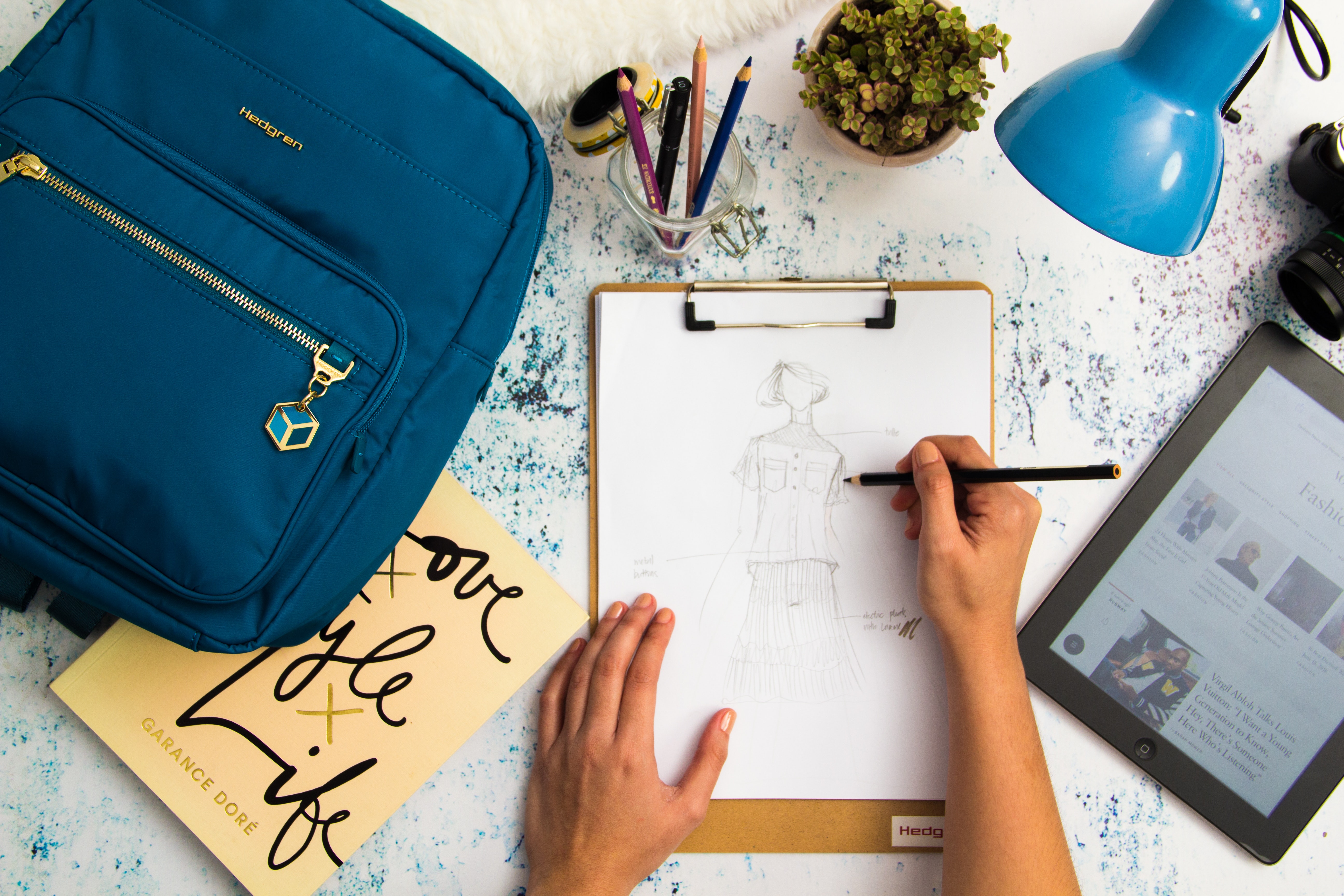 Escritorio con una mochila azul, un flexo azul, una agenda y unas manos diseñando un patrón de moda sobre un folio.