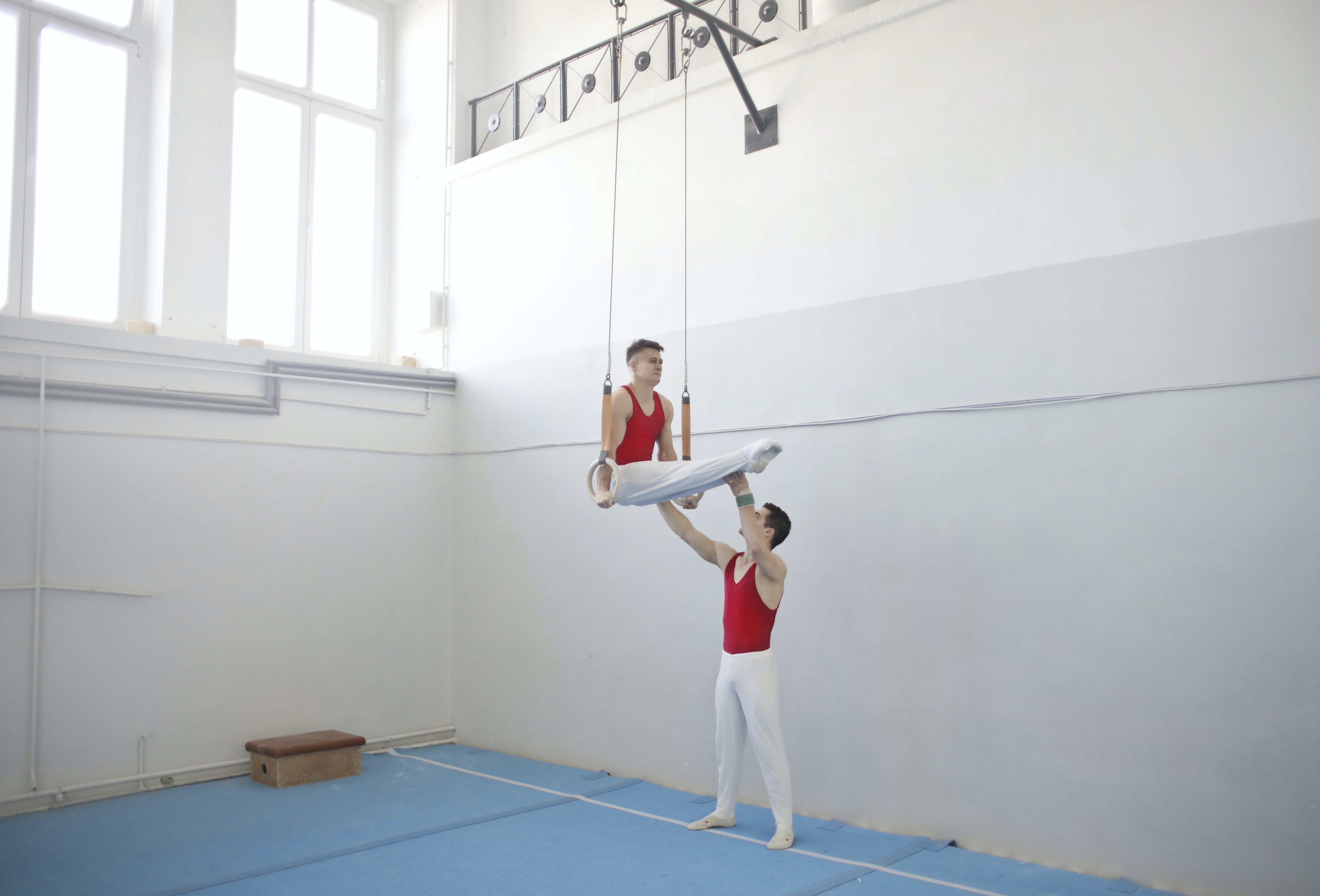 En la imagen se muestran dos atletas de rojo y blanco entrenando en un gimnasio, uno de ellos ayudando al segundo a sostenerse agarrado a unos aros.