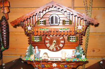 La imagen muestra un reloj de cuco de madera. Simula una casa de madera, con tres personajes a los lados de la puerta principal, un señor mayor a la izquierda con un perro marrón y un niño y una niña con una cabrita blanca a la derecha
