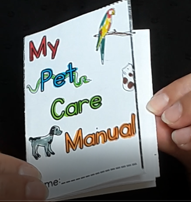 En la fotografía se muestran unas manos abriendo las páginas de un pequeño manual de papel con una portada de mascotas dibujadas y el título My pet care manual.