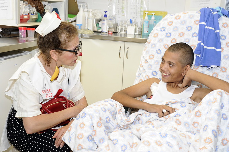 La imagen muestra a una payasa disfrazada de enfermera al costado de la cama en la que un niño la observa sonriendo. 