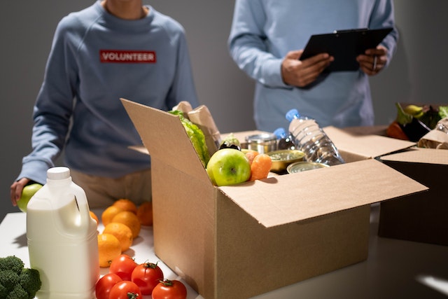 La imagen muestra unas cajas de cartón llenas de comida con voluntarios detrás.