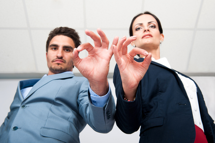 La imagen muestra a dos personas vestidas elegantemente, mirando a cámara y haciendo el signo de OK con las manos.