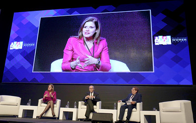 La imagen muestra un escenario en el que hay sentados una mujer y dos hombres y una gran pantalla recoge el gesto de la mujer al hablar.