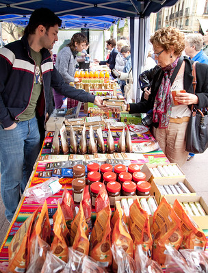 La imagen muestra un puesto de mercado en el que un joven tendero entrega un paquete a una señora. 