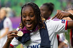 La imagen muestra una deportista que sonríe con euforia y señala la medalla de oro que lleva al cuello.