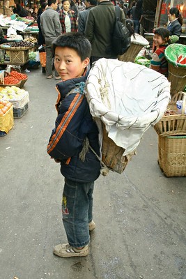 La imagen muestra a un niño en un mercado con una cesta vacía a la espalda.