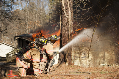 La imagen muestra a dos bomberos que sostienen, el uno tras el otro, una manguera que apuntan hacia un fuego.