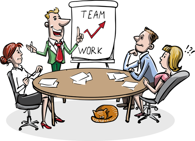  trabajadores sentados alrededor de una mesa redonda atentos a una persona de pie hablando junto a una pizarra en la que se lee team work. 