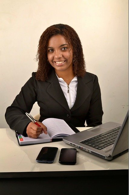 Mujer con traje de chaqueta en una mesa con ordenador portátil, dos móviles y cuaderno con bolígrafo en la mano.