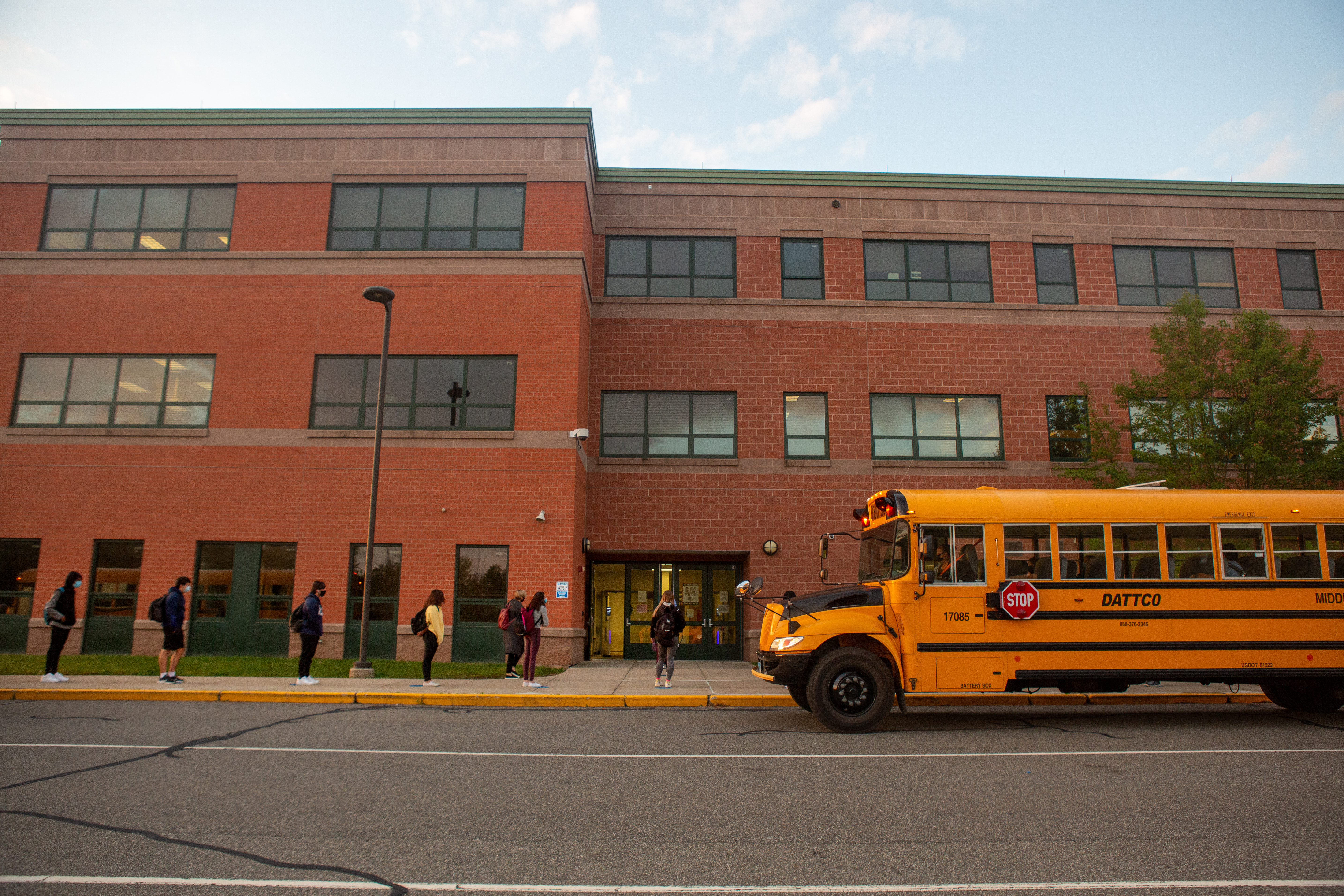 La imagen muestra la fachada de un instituto y un autobús amarillo delante y estudiantes entrando al edificio.