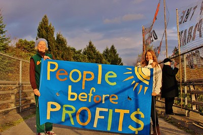 La imagen muestra a dos personas sujetando un cartel hecho de tela que reza “people before profits”.