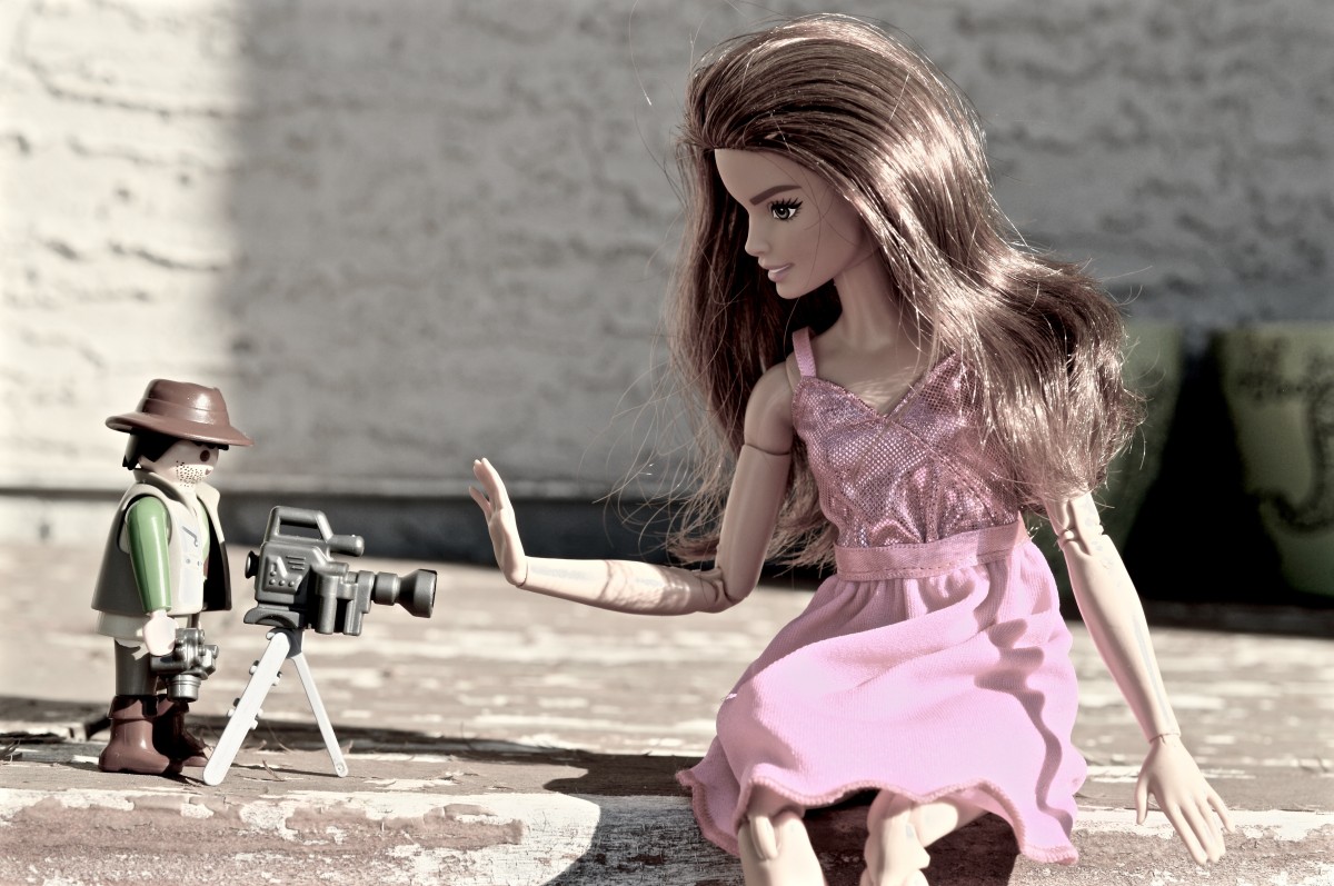 La imagen muestra a una muñeca que está sentada y tapa una cámara de vídeo, detrás de la cámara de video hay un muñeco vestido de reportero.