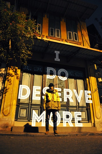 La imagen muestra un chico de pie frente a una fachada en la que se lee ‘I do deserve more’, es decir, me merezco más.