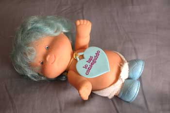 La imagen muestra una muñeca que parece un bebé, con el pelo azul y corto, ojos azules, un pañal blanco y zapatos azules