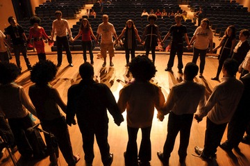 La imagen muestra un teatro. En el escenario hay varios hombres y mujeres cogidos de la mano, haciendo un círculo.