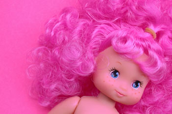 La imagen muestra la cara de una muñeca, con el pelo rosa, rizado y muy largo, sobre un fondo también rosa