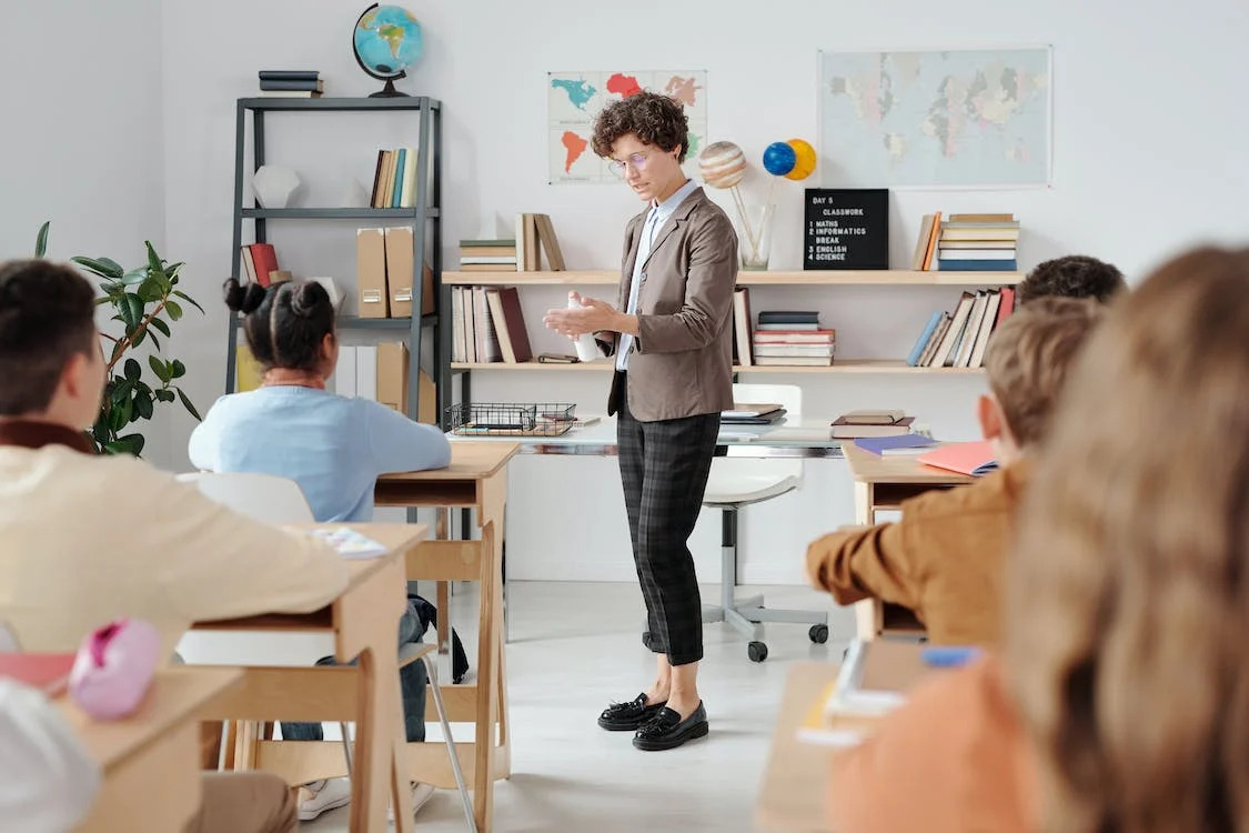 La imagen muestra a una profesora en medio de la clase con los niños y niñas sentados en sus pupitres.