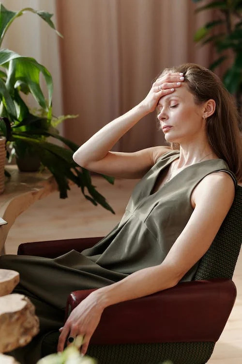 La imagen muestra una mujer sentada en sillón marrón con los ojos cerrados con la mano derecha sobre la frente en señal de dolor. 