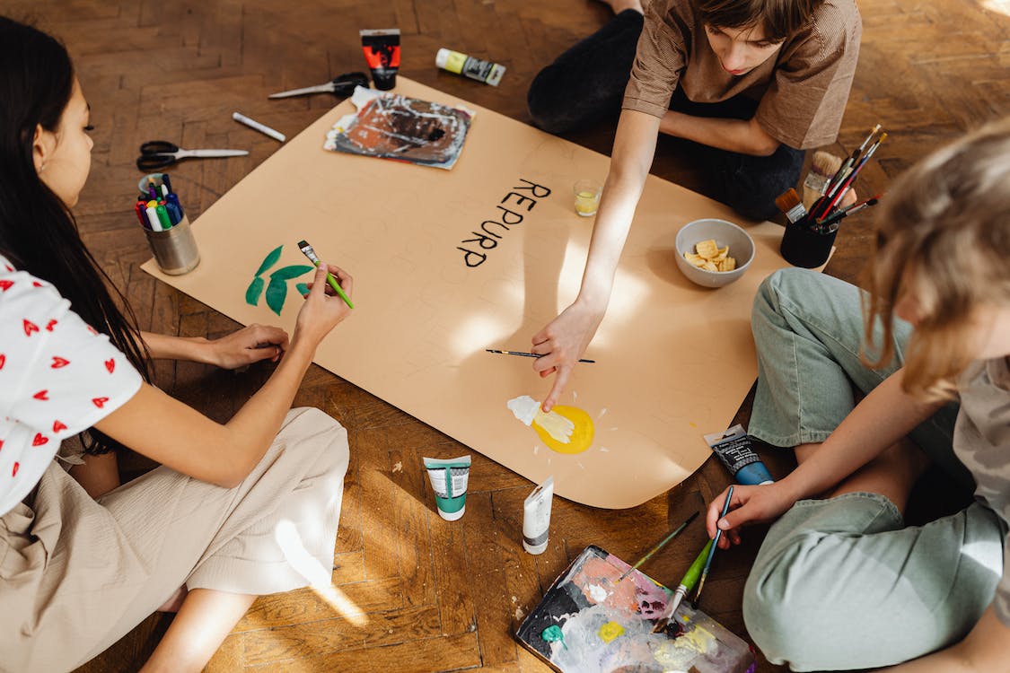 La imagen muestra a tres mujeres sentadas en el suelo diseñando y pintando un póster.