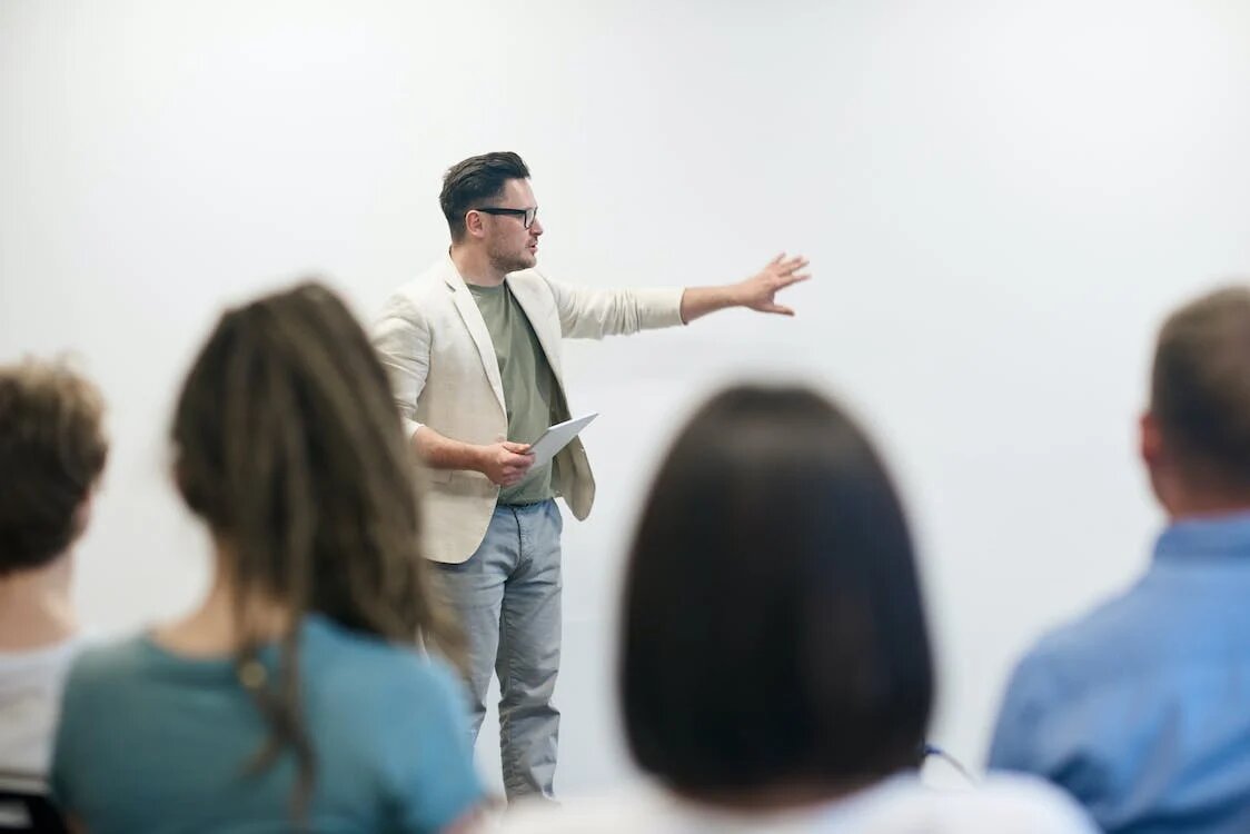 La imagen muestra una clase de estudiantes y al fondo un profesor explicando una actividad a los alumnos y alumnas.