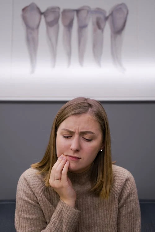 La imagen muestra una mujer que se lleva la mano a la parte baja de la mandíbula. En la pared del fondo hay una imagen de dientes y muelas. 