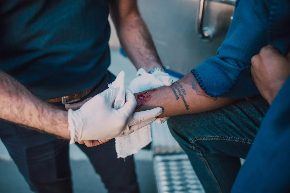 La imagen muestra a un médico curando la herida del brazo de un hombre.