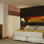 En la imagen se muestra una habitación con una cama de matrimonio entre dos mesitas de noche, dos lámparas de pared y en la pared se ve un cuadro. A la izquierda se ve unas cortinas y parte de un sofá.