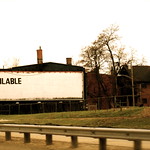 La imagen muestra una valla publicitaria vacía y lista para usarse junto a la carretera y junto a unas viviendas.