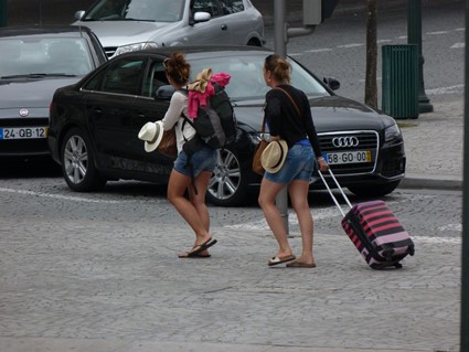 En la imagen se ven dos turistas andando por la calle, una con una mochila grande y otra arrastrando una maleta de ruedas.