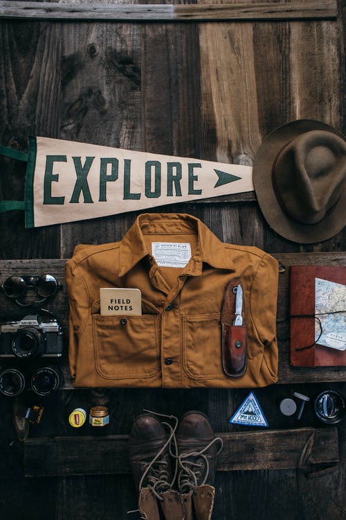 La imagen muestra es un letrero con la palabra explore, una camisa marrón de explorador, un sombrero, unas botas y objetos de exploradores. 