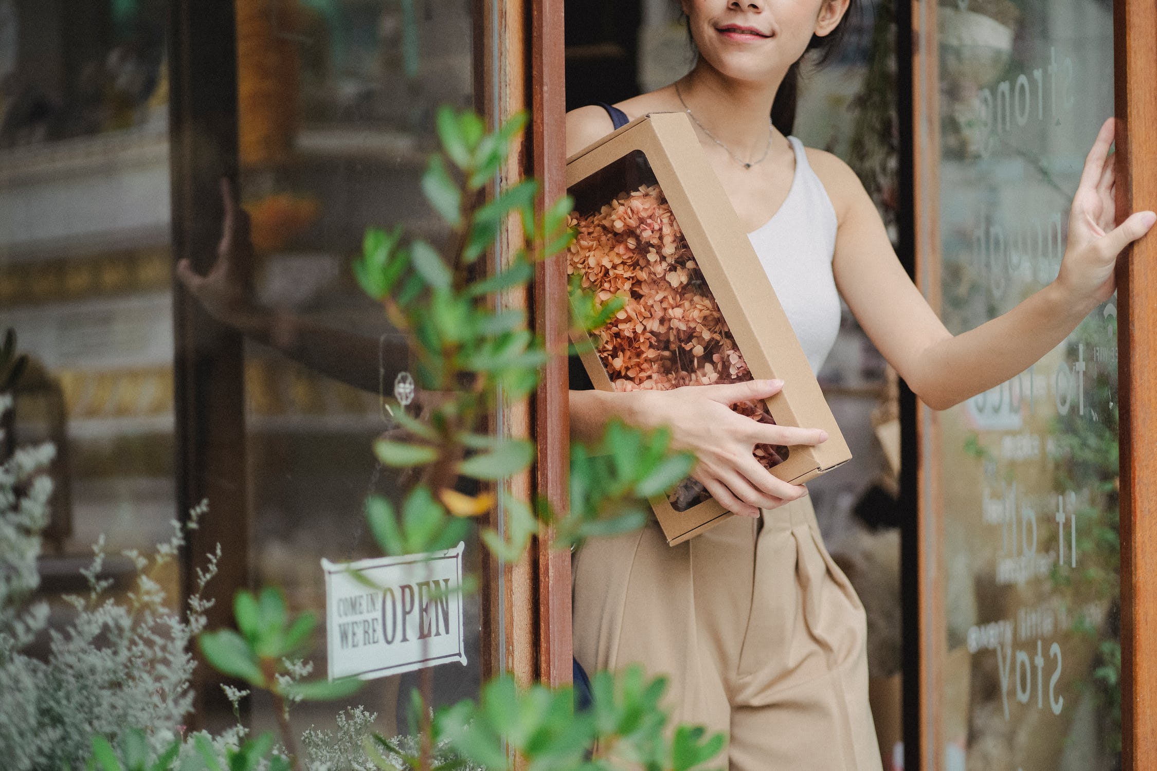 La imagen muestra una mujer con flores secas en la mano saliendo de una floristería abriendo la puerta. 