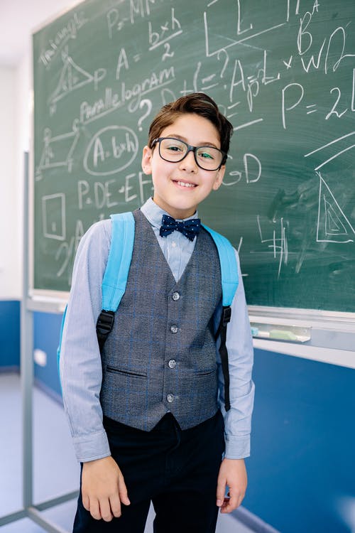 La imagen muestra un niño con gafas redondas, vestido con chaleco azul y pajarita, delante de una pizarra llena de fórmulas matemáticas.