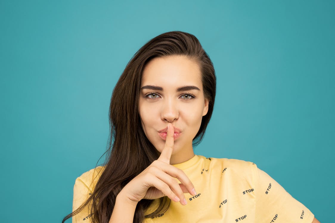 La imagen muestra una mujer con una camiseta amarilla sobre un fondo azul haciendo con su dedo la señal de silencio. 